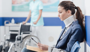 Young female patient with cervical collar support at the hospital, she is sitting in the waiting room and connecting with a digital tablet, medical staff working on the background